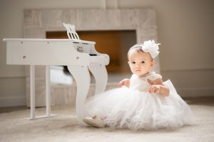 Layla one year old photoshoot wearing white dress sitting beside white kiddie piano