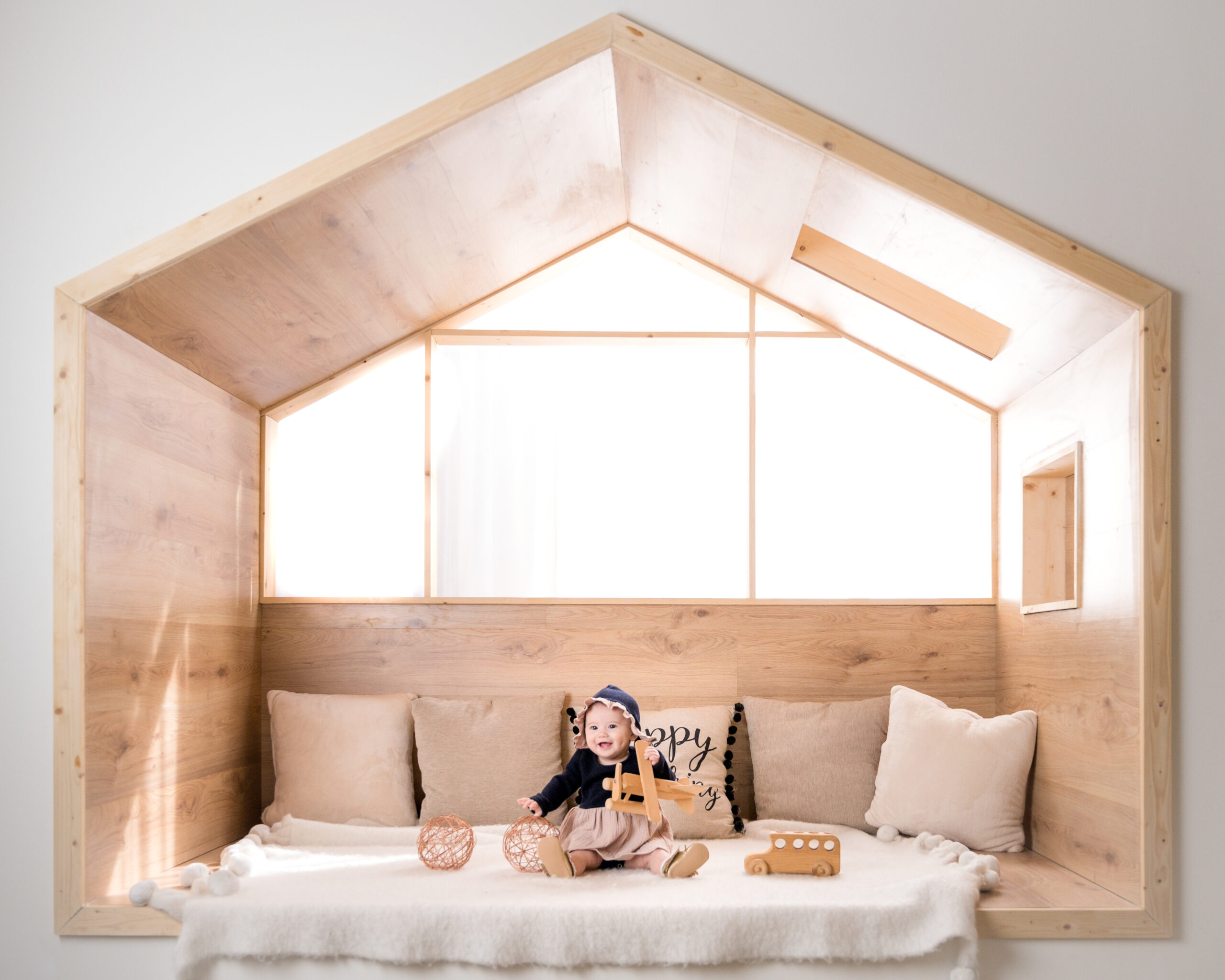 Smiling baby sitting in a cozy wooden nook, playing with wooden toys, captured in soft natural light at gugwa studio